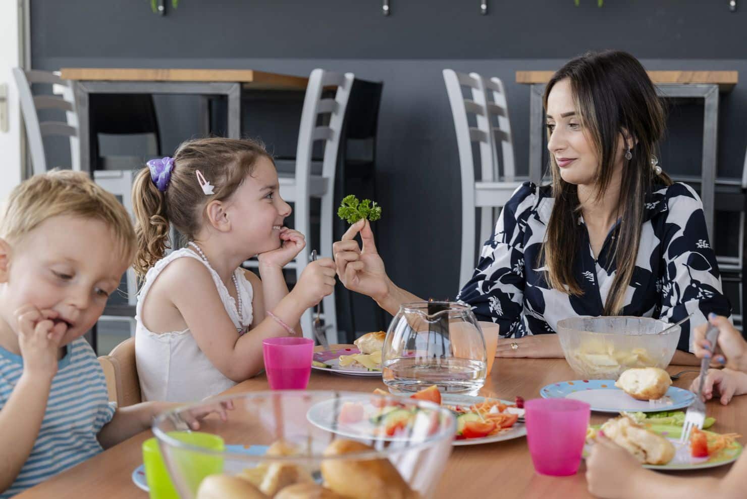 Eine Frau und zwei Kinder sitzen an einem Esstisch mit verschiedenen Lebensmitteln. Die Frau bietet dem einen Kind ein Stück grünes Gemüse an, während das andere Kind isst. Eine Betreuerin der Kinderkrippe miKaDo sitzt mit zwei Kindern an einem Esstisch mit verschiedenen Lebensmitteln. Sie bietet dem Mädchen ein kleinen Stengel Petersilie an , während der Junge im Vordergrund mit den Händen isst.