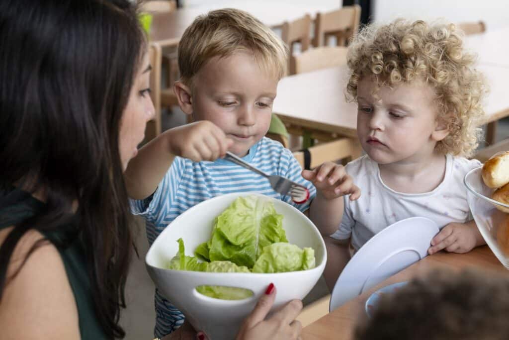 Eine Betreuerin der Kinderkrippe miKaDo und zwei kleine Kinder sitzen an einem Tisch; ein Kind nimmt mit einer Gabel den Salat aus einer Schüssel, während das andere Kind zuschaut.