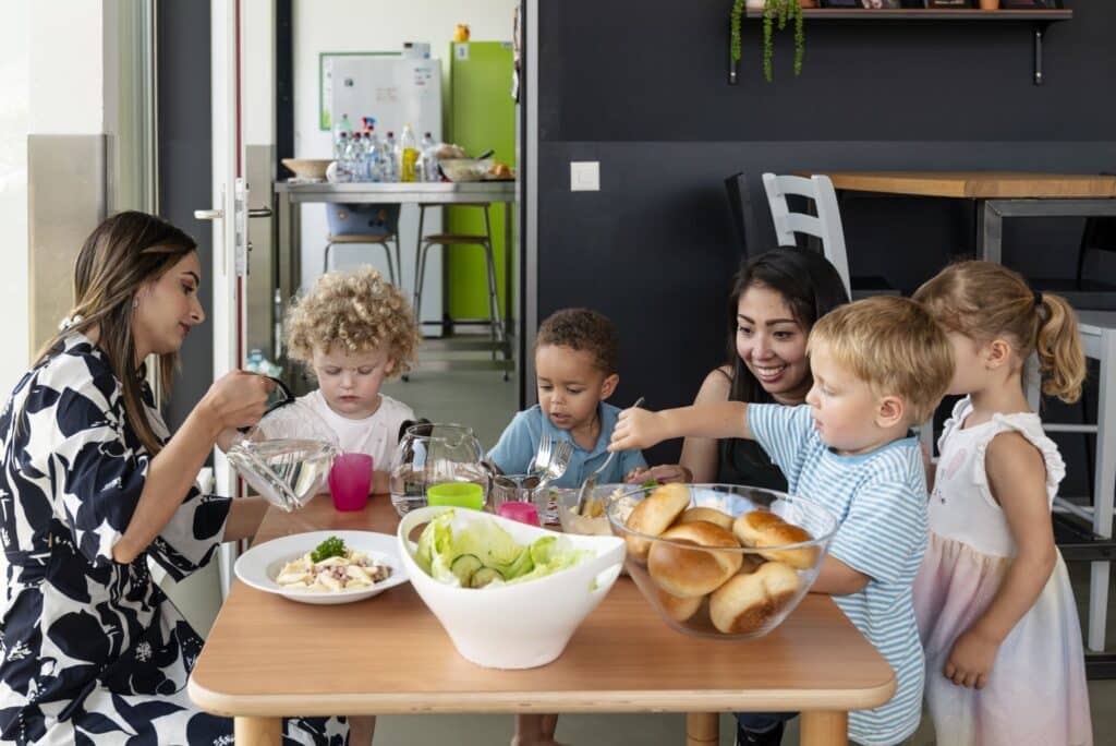 Zwei Betreuerinnen der Kinderkrippe miKaDo und vier kleine Kinder sitzen um einen Tisch mit Schüsseln mit Brot, Salat und anderen Speisen und teilen sich eine Mahlzeit in einer hellen, modernen Küche.