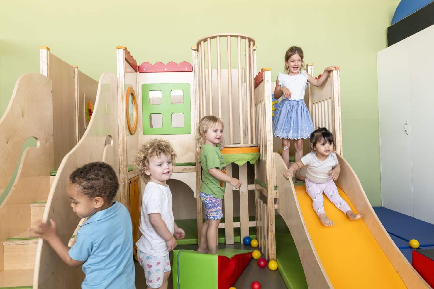 Kinder spielen auf einem Indoor-Klettergrüst mit Rutsche Fünf kleine Kinder spielen auf einem Indoor-Klettergerüst aus Holz mit Rutsche. Einige Kinder stehen unten und beobachten die beiden, die herunterrutschen.