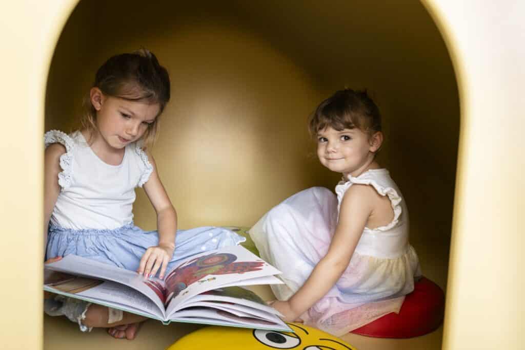 Zwei kleine Mädchen sitzen in einem kleinen geschlossenen Holzraum, der Teil eines Klettergerüst ist. Die eine liest ein Bilderbuch, während die andere daneben sitzt und in die Kamera schaut.