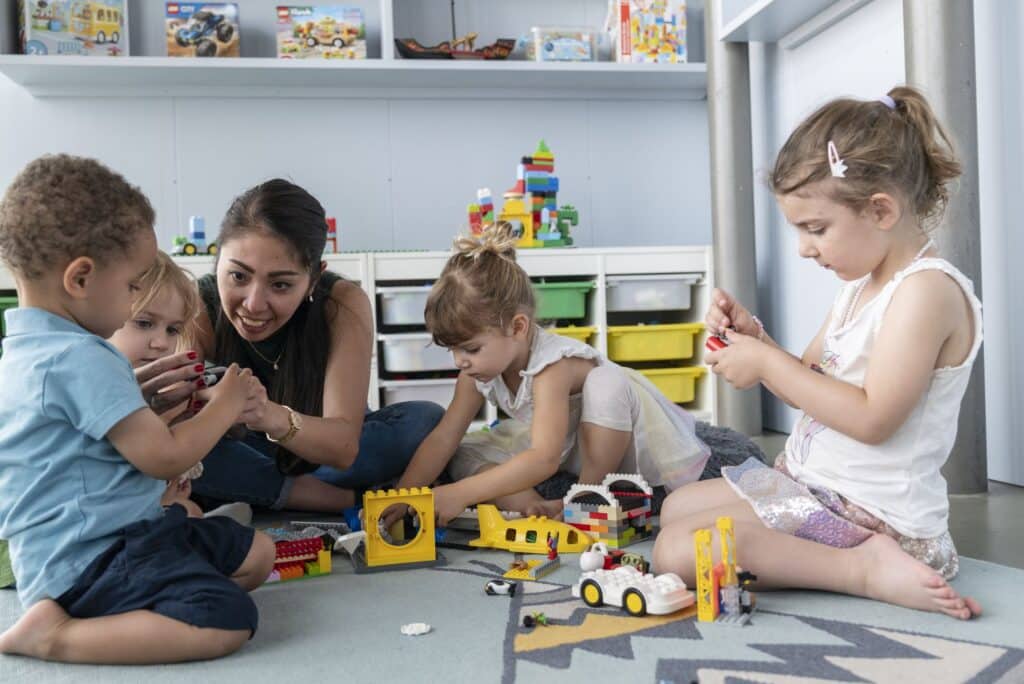 Vier kleine Kinder und eine Betreuerin der Kinderkrippe miKaDo sitzen auf dem Boden und spielen mit Lego in einem Spielzimmer.