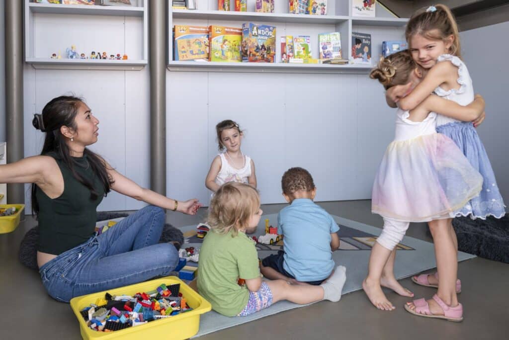 Eine Betreuerin der Kinderkrippe miKaDo sitzt mit vier Kindern auf dem Boden vor einem Regal mit Büchern und Spielen an der Wand und spielt mit Bauklötzen, während rechts zwei Mädchen stehen und sich umarmen.