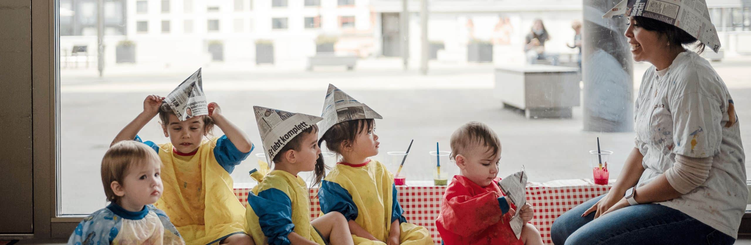 Rechts sitzt eine Betreuerin der Kinderkrippe Mikade mit einem Hut aus Zeitungspapier und lächelt die Gruppe von 5 Kleinkindern an, die vor ihr zum teil ebenfalls mit Hüten aus Zeitungspapier sitzen. Die Kinder haben zum Schutz ihrer Kleidung Hemden aus gelben und blauen Plastik überzogen