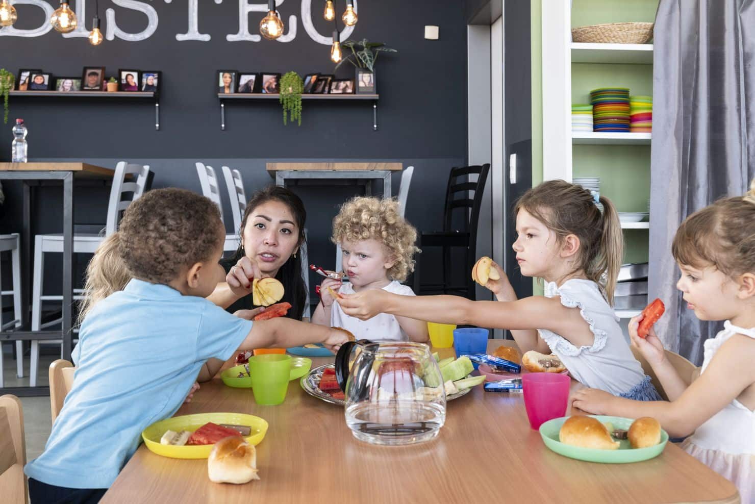 Beim Frühstück in der Kinderkrippe miKaDo Eine Betreuerin der Kinderkrippe miKaDo sitzt mit fünf Kindern an einem Tisch und isst Obst. Gemüse und Brotchen von buntem Plastikgeschirr. Ein Mädchen reicht dem Kinde gegenüber ein Stück Paprika.