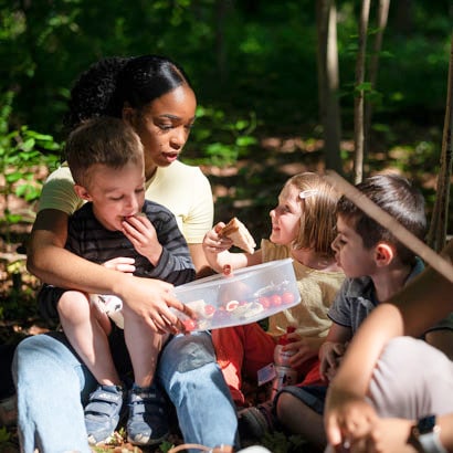 Farbige Betreuerin der Kinderkrippe miKaDo sitzt umringt von Kindern im Wald und hält eine Frischhaltedose mit Tomaten und Brotstücken in der Hand und alle essen davon