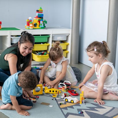 Betreuerin der Kinderkrippe miKaDo sitzt mit Kindern auf einem Spielteppisch und alle spielen mit Lego