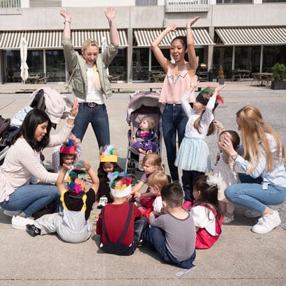 Kinder sitzen und stehen mit vier Betreuerinnen der Kinderkrippe miKaDo im Kreis, singen und heben dabei die Hände in die Luft
