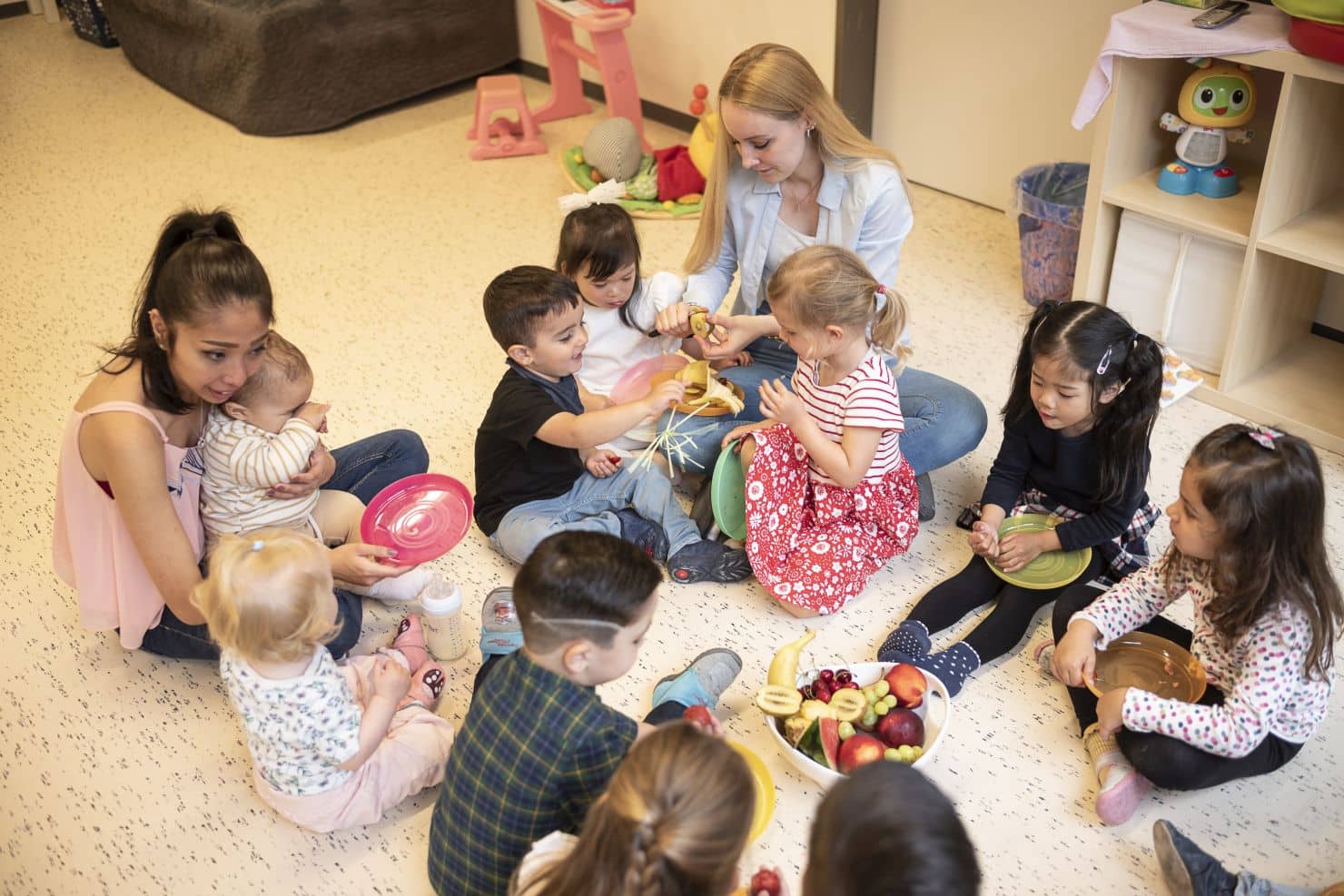 Zwei Betreuerinnen der Kinderkrippe miKaDo sitzen mit einer Gruppe Kinder im Kreis auf dem Boden und essen vorbereitetes Obst beim Zvieri.