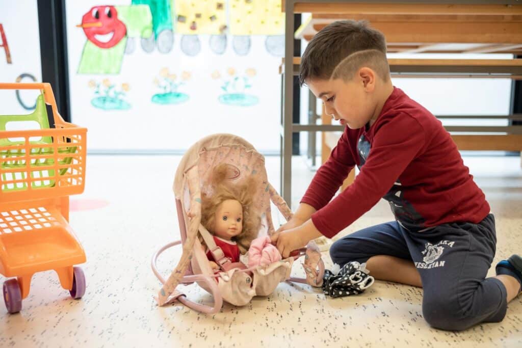 Ein Junge in einem roten Pullover kniet auf dem Boden und setzt eine Puppe in einen Spielzeug-Babysitz. Daneben steht ein Spielzeug-Einkaufswagen.