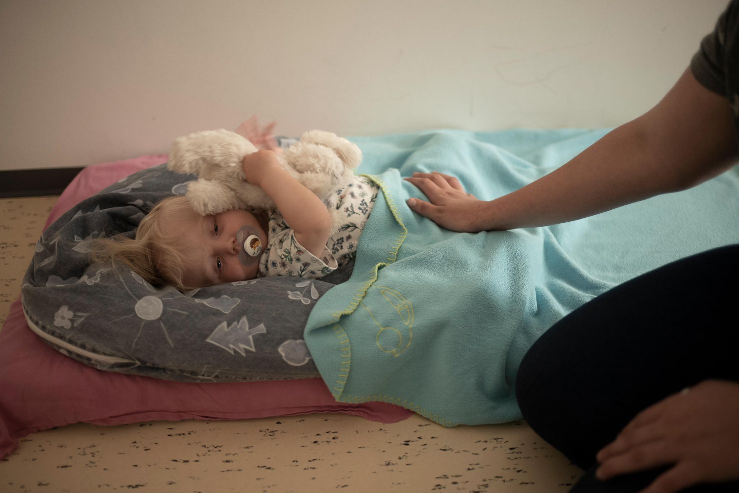 Ein Kleinkind liegt mit einem Schnuller auf einer Matte, umarmt ein Stofftier und ist mit einer Decke zugedeckt, während die Hand der Betreuerin der Kinderkrippe miKaDo sanft auf seinem Bauch ruht.