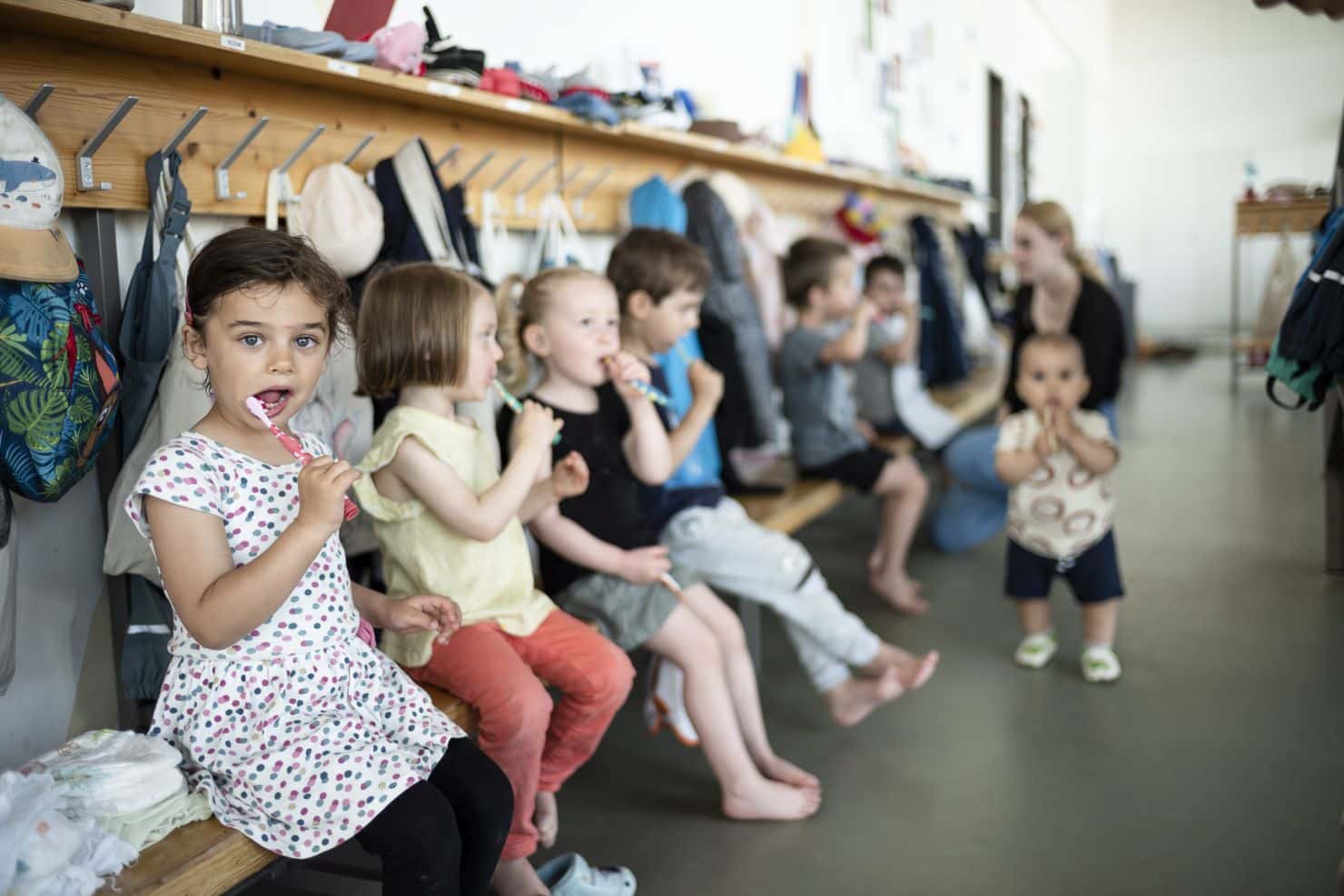 Mehrere kleine Kinder sitzen in einer Reihe auf einer Bank und putzen sich die Zähne. Über der Bank befinden sich Haken an der Wand mit Kleidungsstücken und Taschen. Im Hintergund steht ein Kleinkind und dahinter eine Betreuerin der Kinderkrippe miKaDo, die mit einem Jungen spricht, der sich ebenfalls die Zähne putzt.