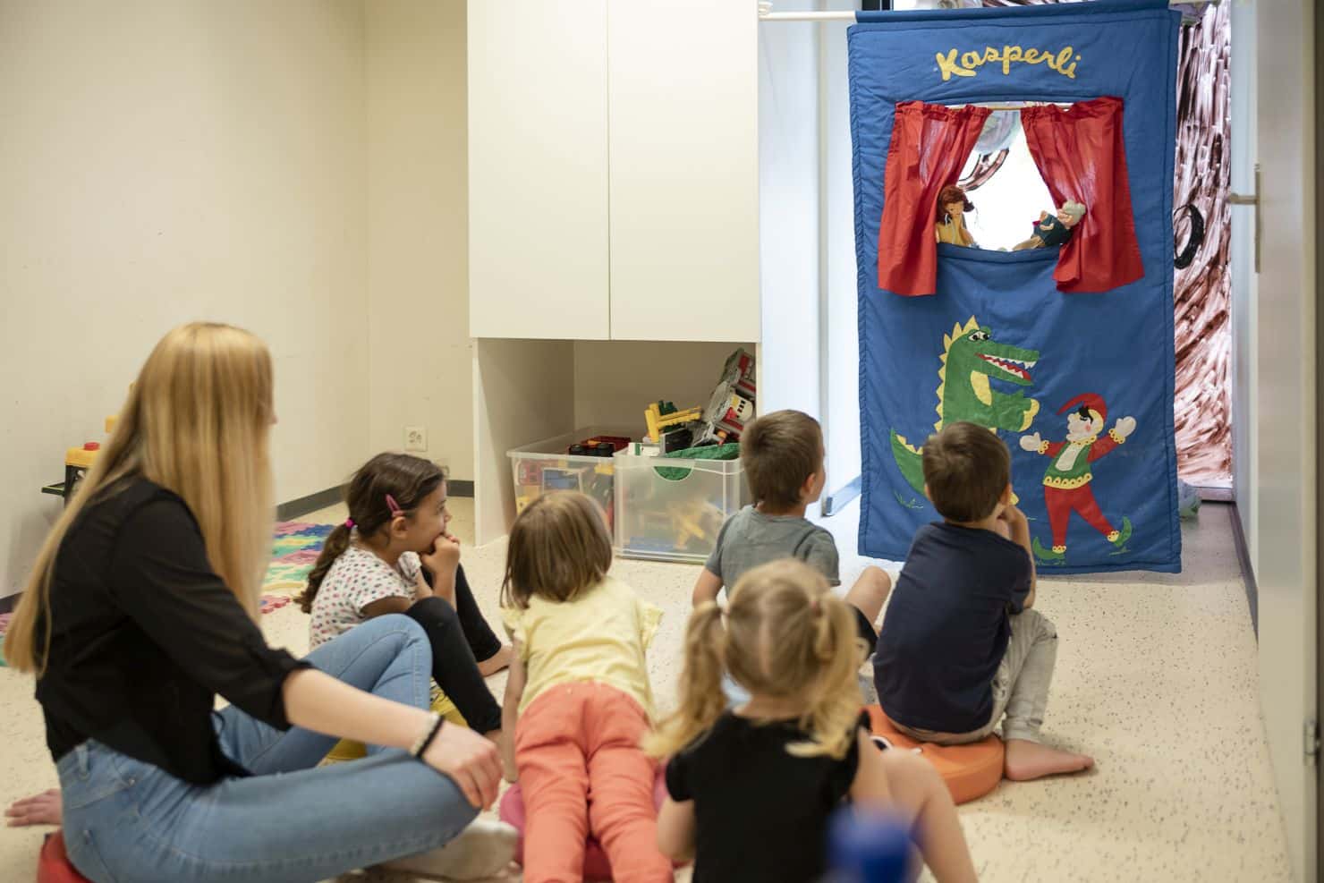Eine Gruppe von Kindern und eine Betreuerin der Kinderkrippe miKaDositzen auf dem Boden und beobachten eine Kasperli-Vorstellung.