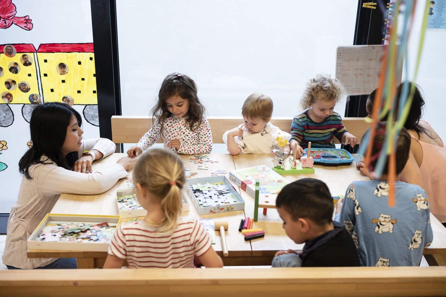 Eine Gruppe kleiner Kinder sitzen um einen Tisch. Sie setzen Puzzles zusammen oder setzen Baukästen zusammen. Im Hintergrund bunt geschmückte Fenster.