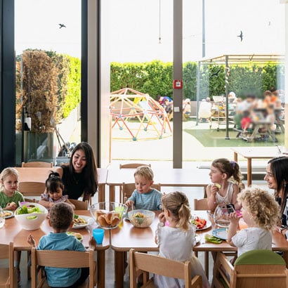 Eine Gruppe kleiner Kinder sitzt an Tischen und isst in einem hellen, modernen Raum mit großen Fenstern und Blick nach draußen, Zwischen ihnen sitzen zwei Betreuerinnen der Kinderkrippe miKaDo. Im Hintergund sieht man draussen weitere Kindergruppen im Grünen sitzen und essen.