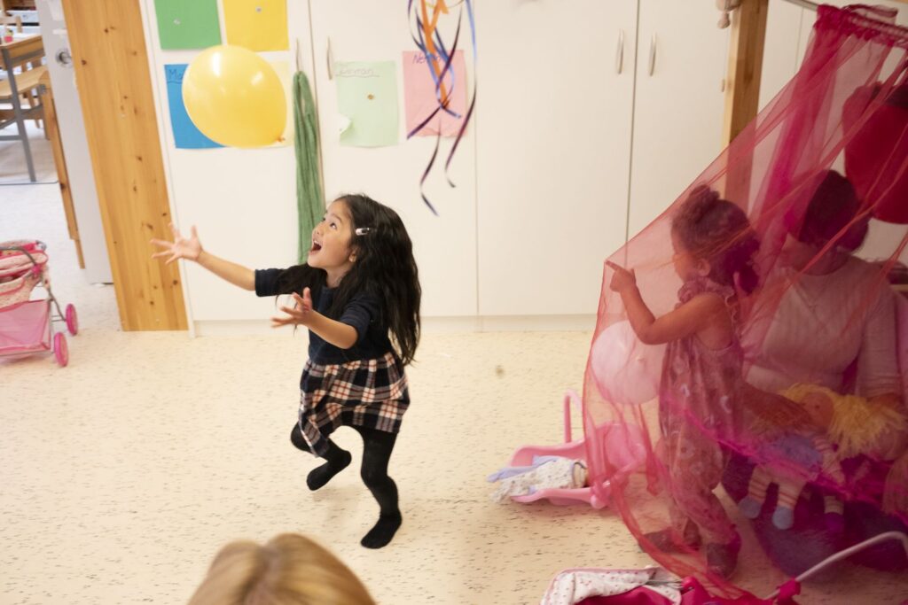Ein kleines Mädchen springt einem gelben Luftballon über ihr nach während andere Kinder unter einem rosa Baldachin in einem Spielzimmer spielen.