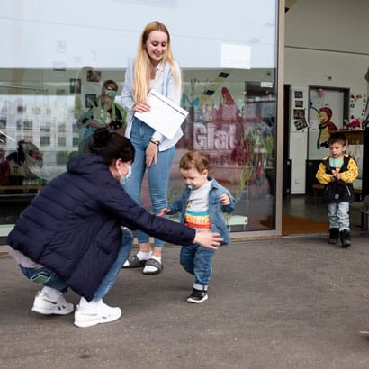 Kind kommt aus der Kinderkrippe miKaDo und Mutter beugt sich mit ausgebreiteten Armen zur Begruessung herunter.