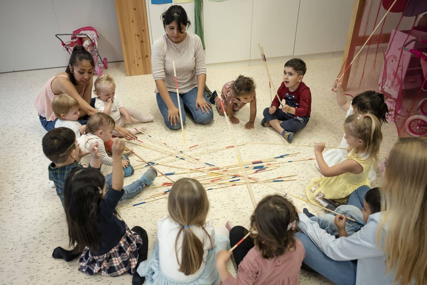 Eine Gruppe kleiner Kinder und zwei Betreuerinnen der Kinderkrippe miKaDo sitzen in einem Kreis auf dem Boden und spielen ein grosses Mikado. Die langen Mikadostäbe liegen in der Mitte verstreut.