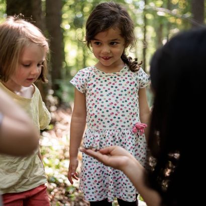 Zwei kleine Mädchen stehen in einer Gruppe im Wald und hören den Erklärungen der Betreuerin aus der Kinderkrippe Mikado so
