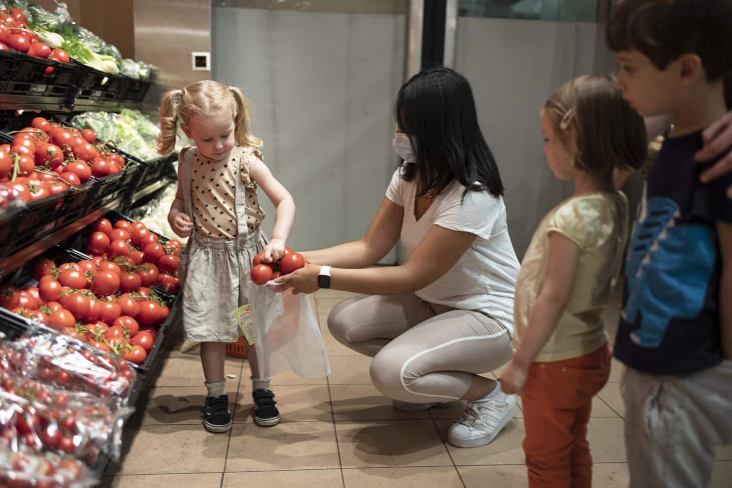 In der Gemüseabteilung eines Lebensmittelgeschäfts legt ein Mädchen Tomaten in einen Einkaufssack, den ihr die Betreuerin der Kinderkrippe miKaDo hin hält während zwei andere Kinder zusehen.
