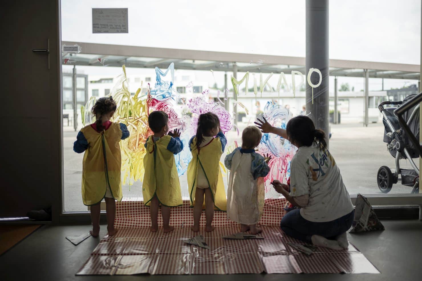 Vier kleine Kinder mit Schutzkittel aus Plastik stehen am Femster und bemalen es mit bunten Mustern. Die Betreuerin der Kinderkrippe miKaDo hockt neben ihnen und malt mit. Rechts sieht man draußen vor dem Fenster noch einen Kinderwagen stehen.