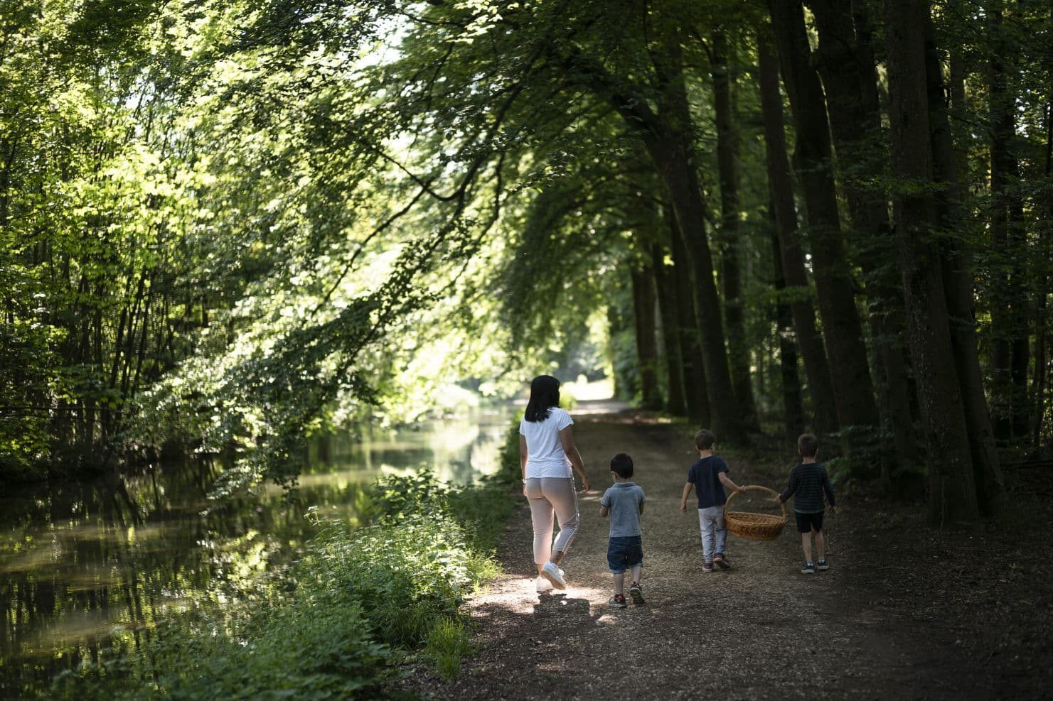 Eine Betreuerin der Kinderrkrippe miKaDo und drei Kinder gehen auf einem Waldweg an einem ruhigen Fluss entlang. Die Kinder tragen einen großen Korb. Das Sonnenlicht fällt durch die Bäume.