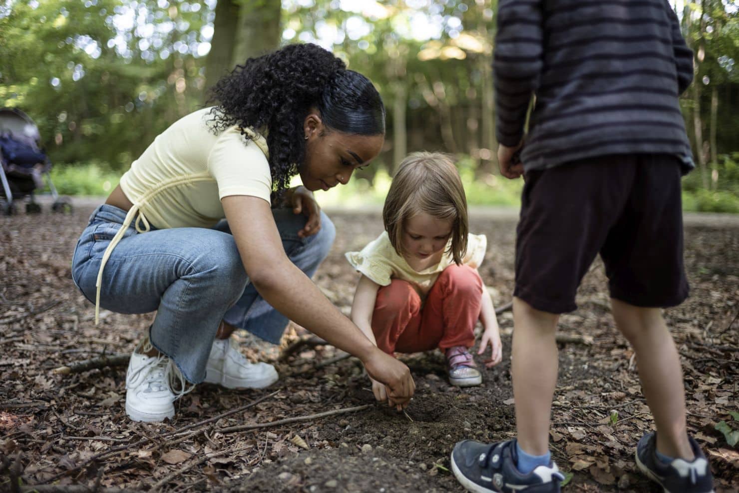 Eine Betreuerin der Kinderkrippe miKaDo und zwei Kinder kauern auf dem Waldboden und untersuchen gemeinsam den Boden zwischen Bäumen und verstreuten Blättern.