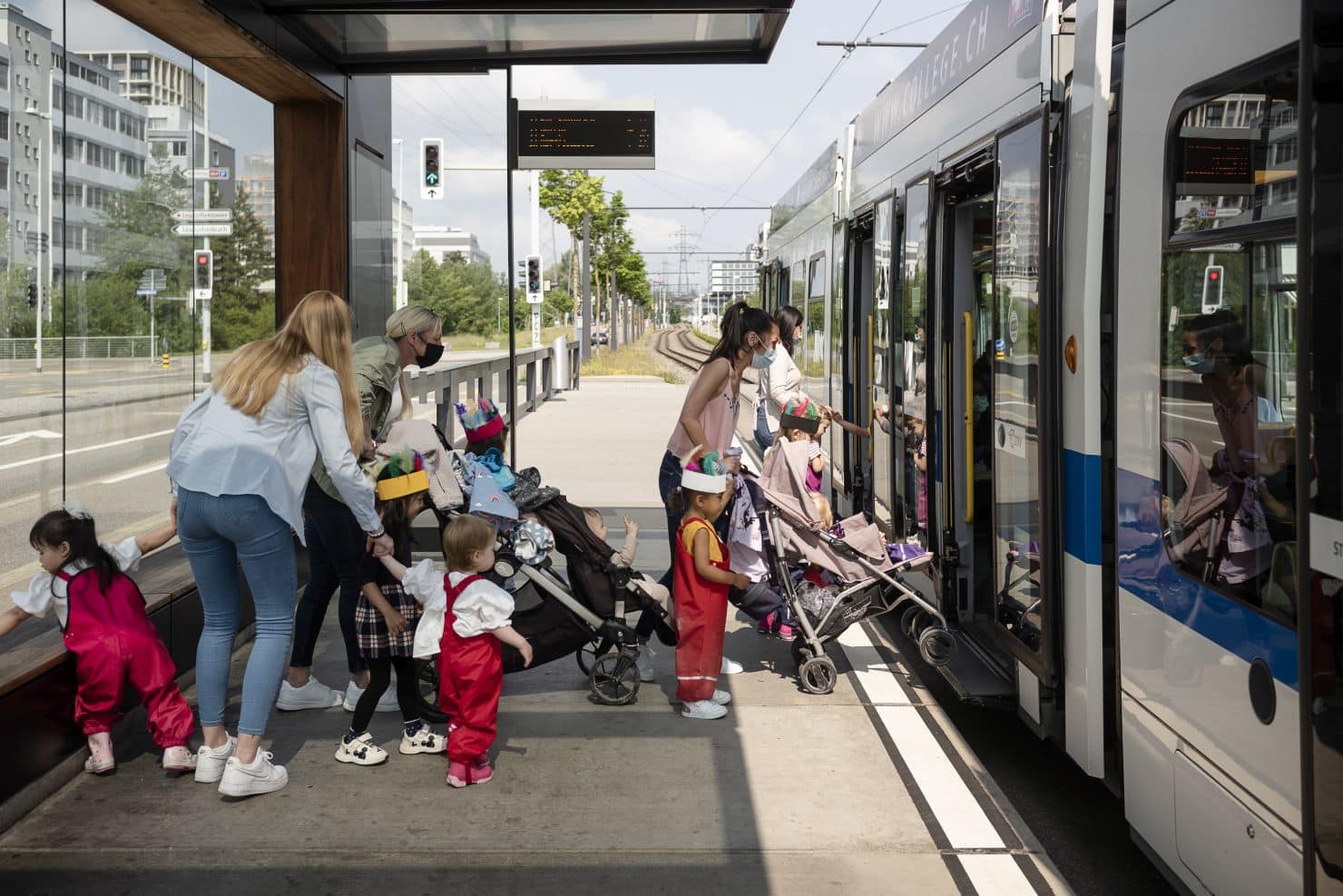 Mehrere Betreuerinnen der Kinderkrippe miKaDo steigen mit kleinen Kindern und Kinderwagen an einem sonnigen Tag an einer Haltestelle in der Stadt in eine Tram ein.