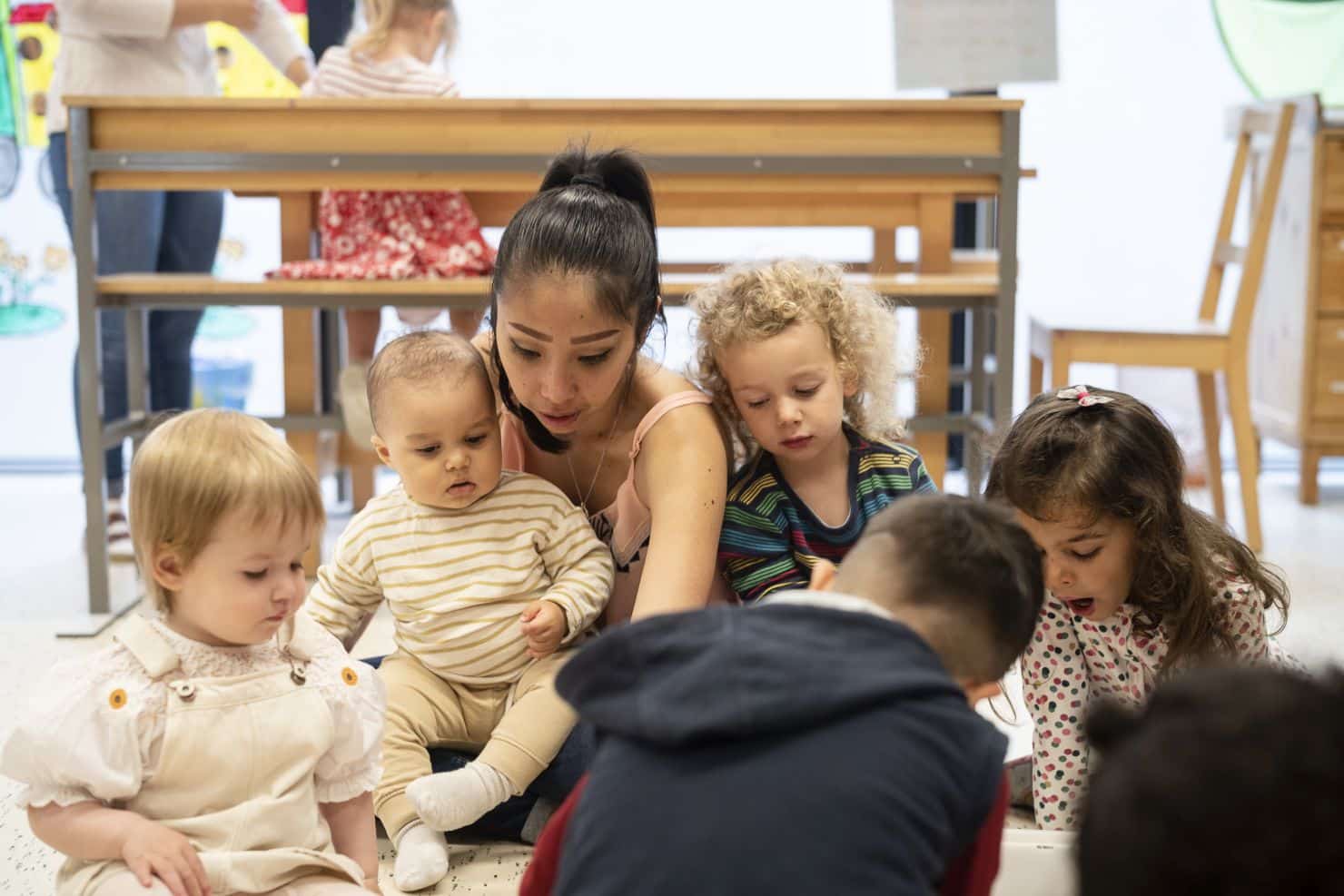 Eine Betreuerin der Kinderkrippe miKaDo sitzt mit einer Gruppe kleiner Kinder auf dem Boden im Morgenkreis und schauen auf etwas, was in der Mitte vor ihnen liegt.