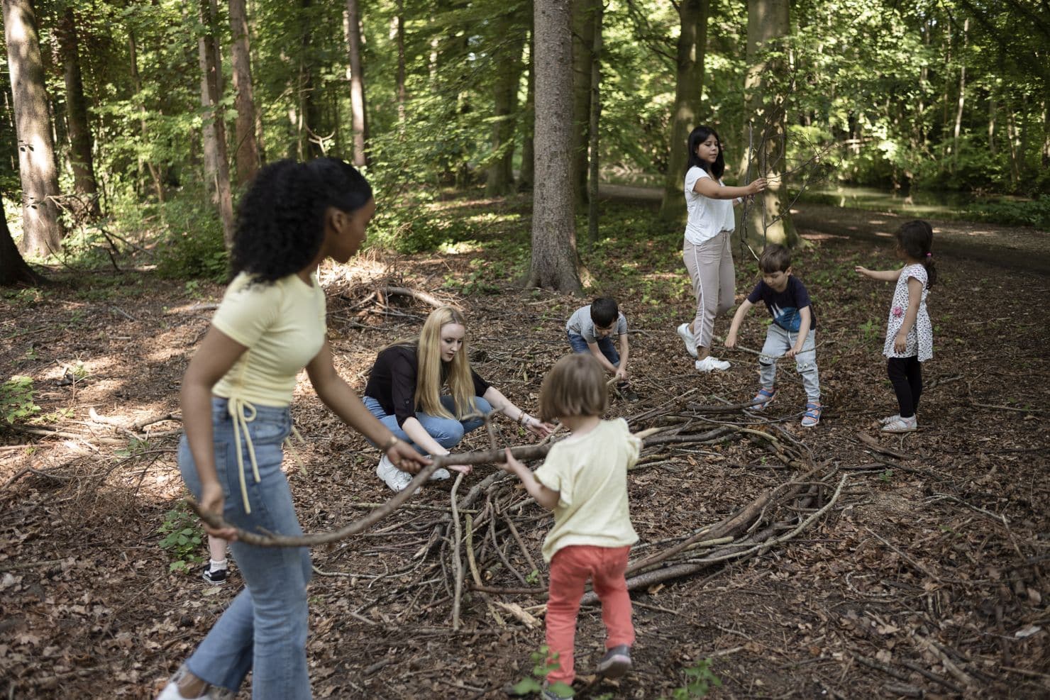 Eine Gruppe von Kindern und zwei Betreuerinnen der Kinderkrippe miKaDo legen auf einer sonnenbeschienenen Waldlichtung Stöcke in einem Kreis auf den Boden.