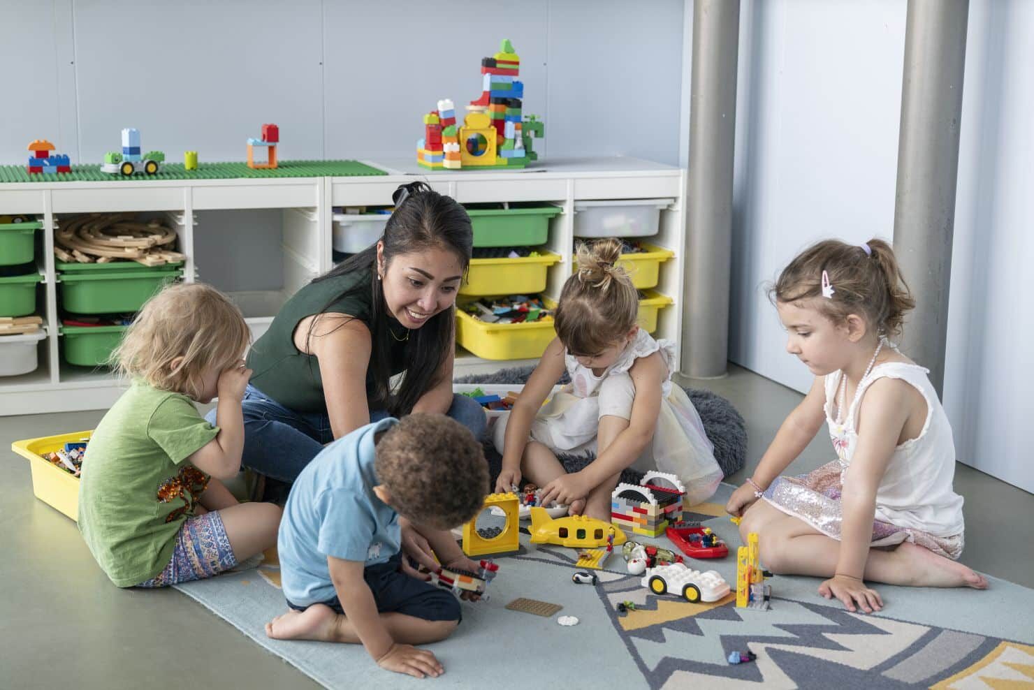 Eine Betreuerin und vier kleine Kinder sitzen in einem Spielzimmer auf dem Boden und spielen mit verschiedenen Bausteinen und Spielzeugfahrzeugen. Im Hintergrund sind Regale mit organisiertem Spielzeug zu sehen. Eine Betreuerin der Kinderkrippe miKaDo und vier kleine Kinder sitzen in einem Spielzimmer auf dem Boden und spielen mit verschiedenen Bausteinen und Spielzeugfahrzeugen. Im Hintergrund sind Regale mit organisiertem Spielzeug zu sehen.