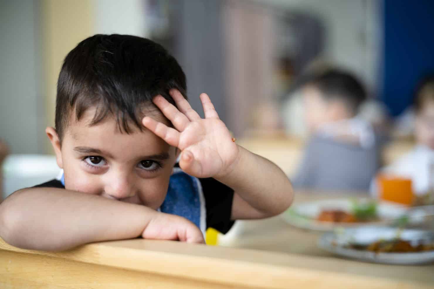 Ein kleiner Junge mit kurzen dunklen Haaren winkt und lehnt sich lächelnd an Rückenlehne der Bank auf der er sitzt. Auf dem Tisch stehen Teller mit Essen, und andere Kinder sind unscharf im Hintergrund zu erkennen.