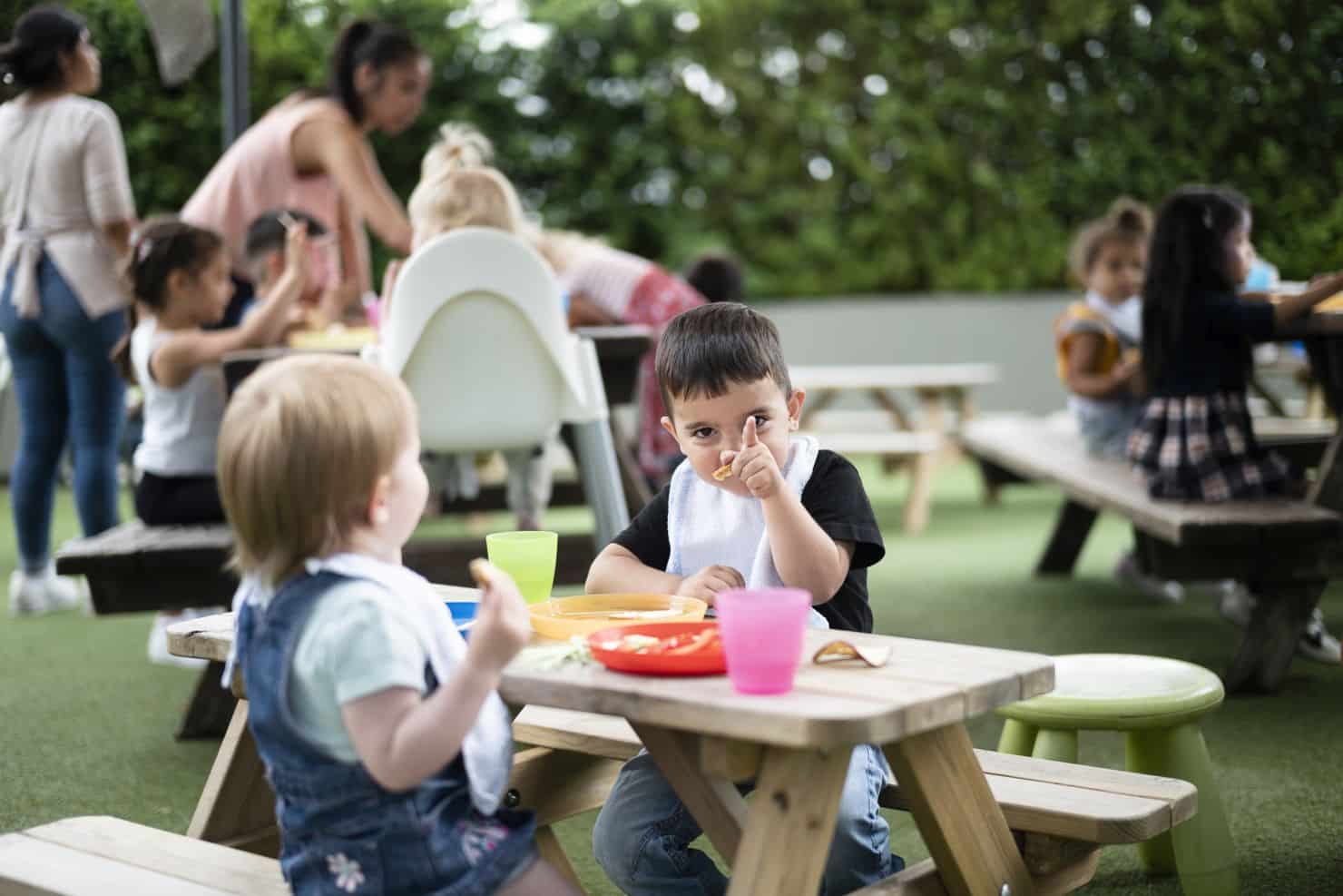 Kinder sitzen an Picknicktischen im Freien und essen Snacks. Ein Junge im Vordergrund zeigt auf den Fotografen, während ein anderes Kind ihm gegenübersitzt. Andere Kinder und Erwachsene sind im Hintergrund zu sehen.