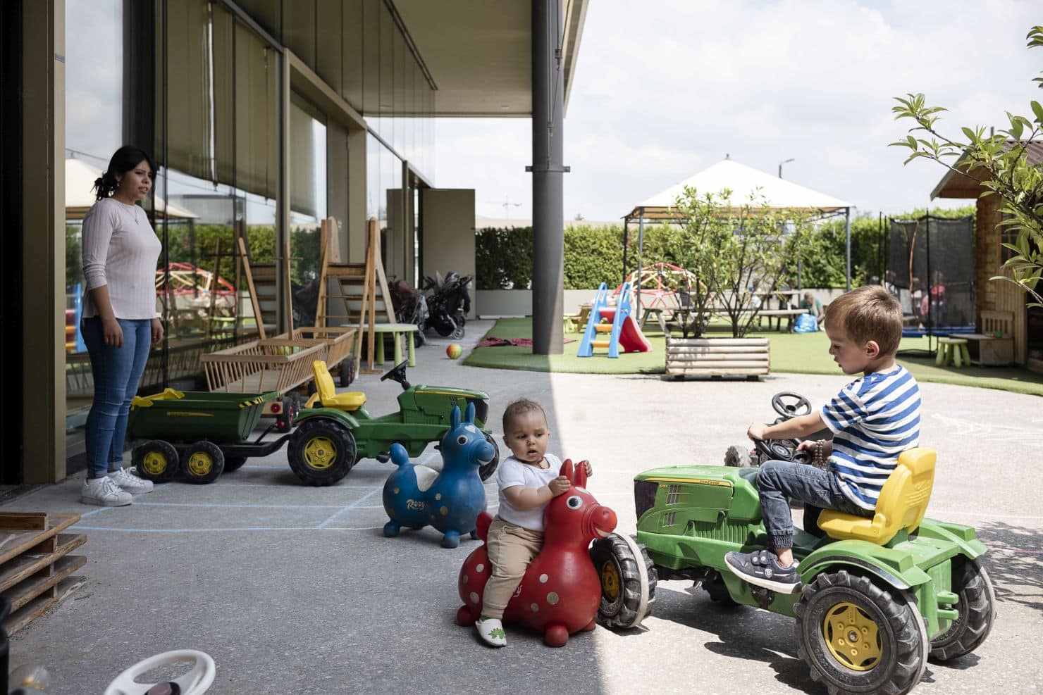 Eine Betreuerin der Kinderkrippe miKaDo steht draußen und beobachtet zwei kleine Kinder, die auf dem Spielplatz der Kinderkrippe mit einem Laufdino und einem Spielzeugtraktor spielen.