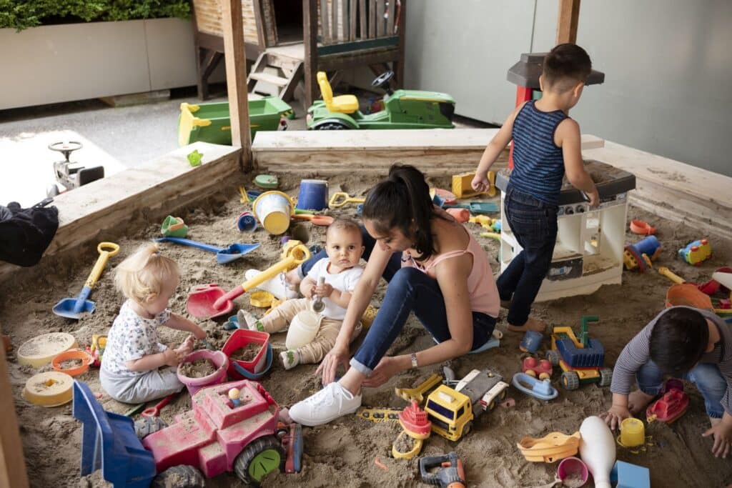 Mehrere kleine Kinder und eine Betreuerin der Kinderkrippe miKaDo spielen in einem Sandkasten, der mit Spielzeug wie Lastwagen, Schaufeln und Eimern gefüllt ist.