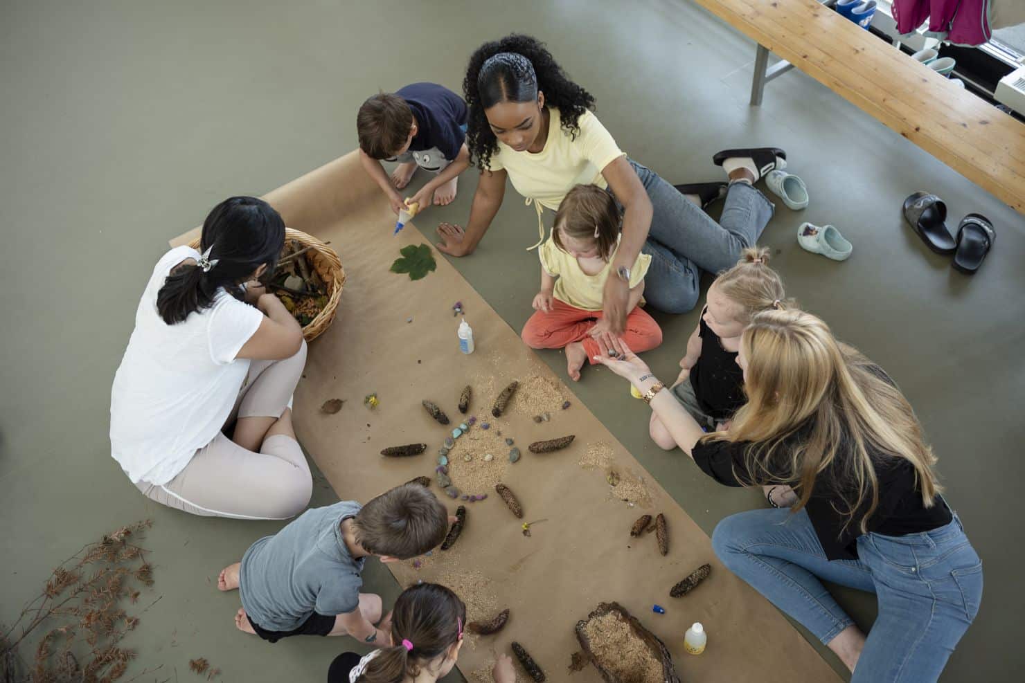 Kinder und Erwachsene sitzen auf dem Boden um ein Blatt Papier herum und ordnen Naturmaterialien wie Blätter, Tannenzapfen und Sand zu Mustern für ein Kunstprojekt an.