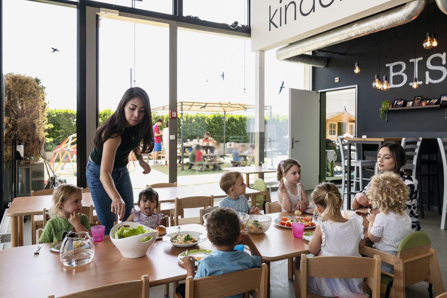 Kinder sitzen mit Betreuerinnen beim Mittagessen Eine Gruppe kleiner Kinder sitzt an Tischen und isst in einem hellen, modernen Raum mit großen Fenstern und Blick nach draußen, Eine Betreuerinnen der Kinderkrippe miKaDo sitzt am Kopfende des Tisches, die andere steht hinter den Kindern und rührt den Salat um. Im Hintergund sieht man draussen weitere Kindergruppen im Grünen sitzen und essen.