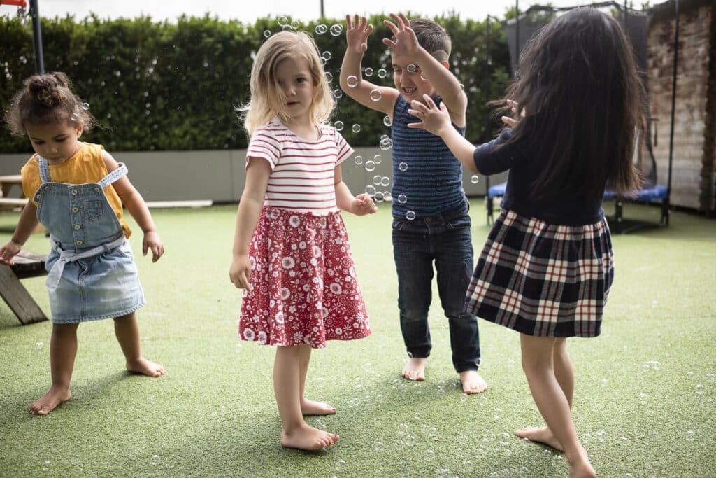 Vier kleine Kinder spielen barfuß auf Kunstrasen und greifen nach Seifenblasen Im Aussenbereich der Kinderkrippe miKaDo.