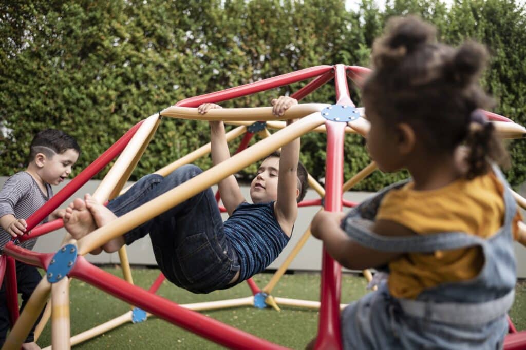 Drei kleine Kinder spielen auf einem rot-gelben Klettergerüst im Freien, wobei ein Kind im Inneren des Geräts hängt und zwei andere in der Nähe klettern oder sitzen.