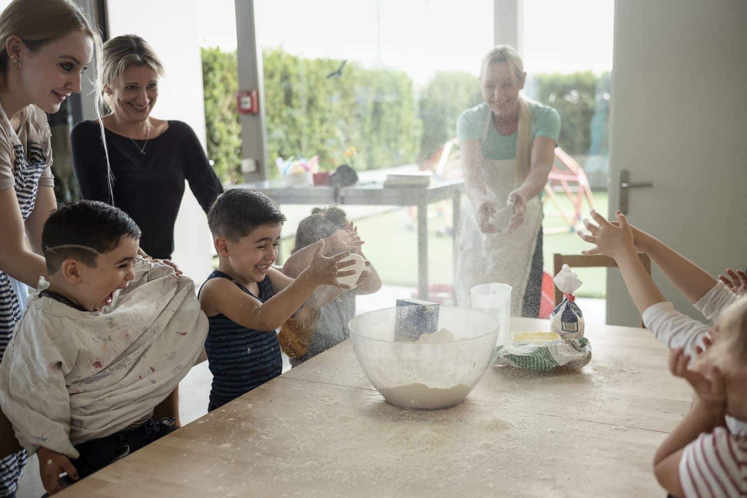 Eine Gruppe von Kindern und drei Betreuerinnen der Kinderkrippe miKaDo stehen um einen Tisch und haben Spass dabei Mehl spielerisch Mehl in die Luft über der Schüssel auf dem Tisch zu werfen. Im Hintergrund sieht man durch ein grosses Fenster in den grünen Garten.