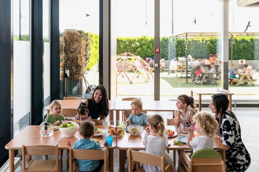 Eine Gruppe kleiner Kinder sitzt an Tischen und isst in einem hellen, modernen Raum mit großen Fenstern und Blick nach draußen, Zwischen ihnen sitzen zwei Betreuerinnen der Kinderkrippe miKaDo. Im Hintergund sieht man draussen weitere Kindergruppen im Grünen sitzen und essen.