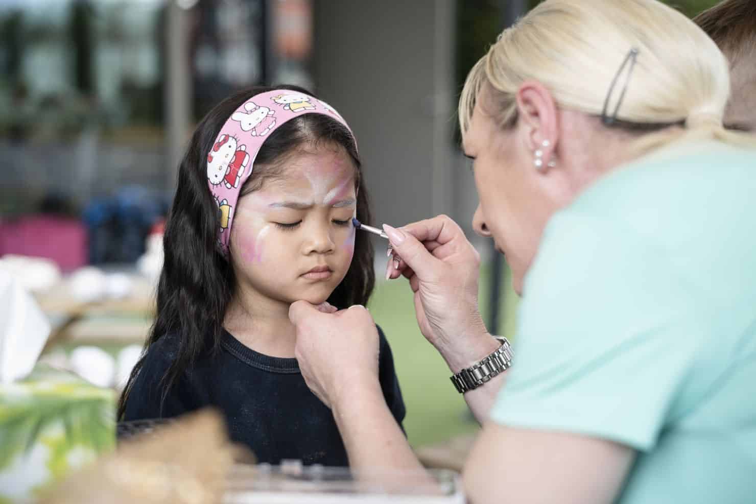 Ein kleines Mädchen mit einem Stirnband lässt sich von einer Betreuerin der Kinderkrippe miKaDo mit einem kleinen Pinsel das Gesicht bemalen. Das Mädchen sitzt dabei still und hat die Augen geschlossen.