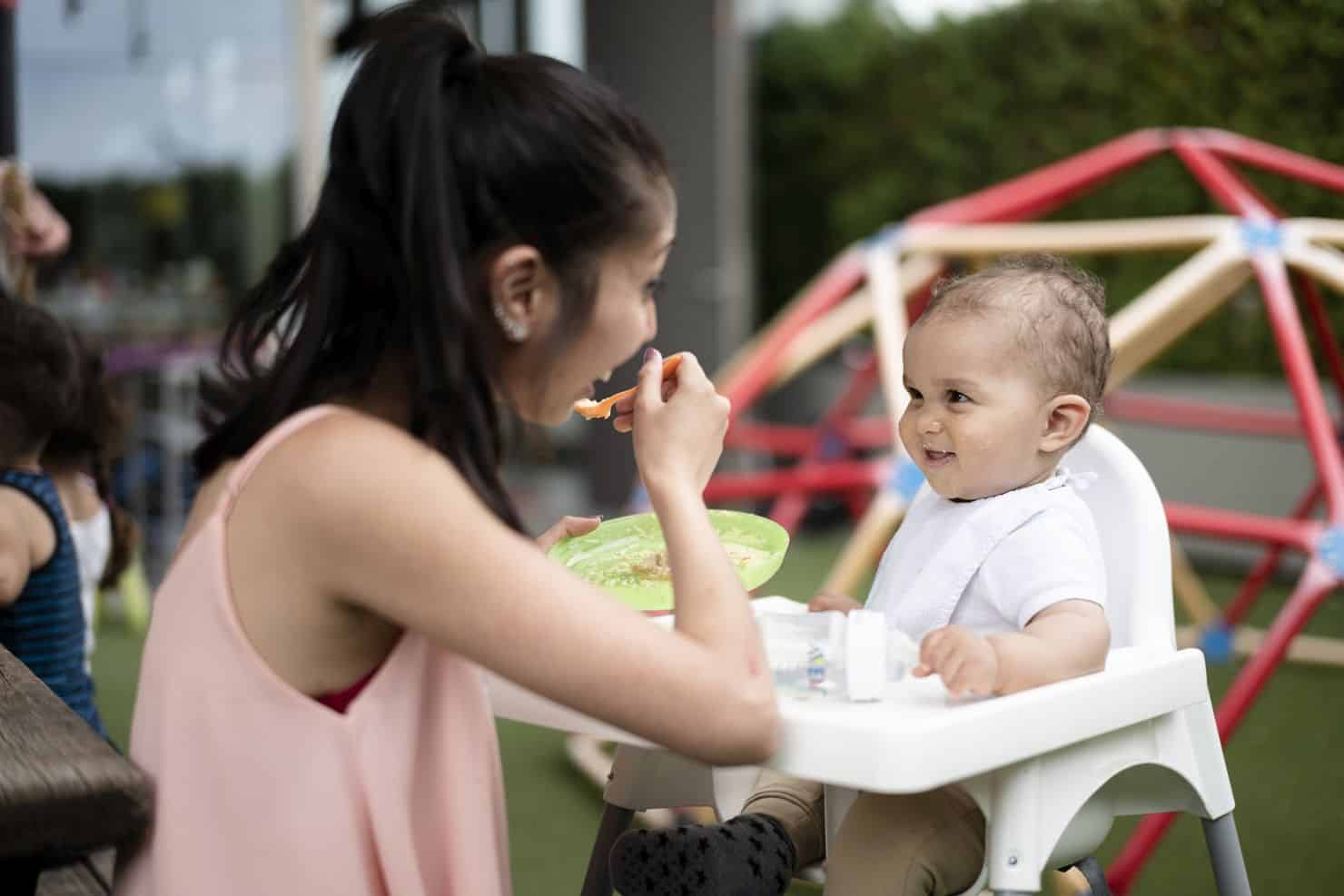 Eine Betreuerin der Kinderkrippe miKaDo füttert ein lächelndes Baby in einem Hochstuhl im Freien. Im Hintergrund sind Spielgeräte zu sehen.