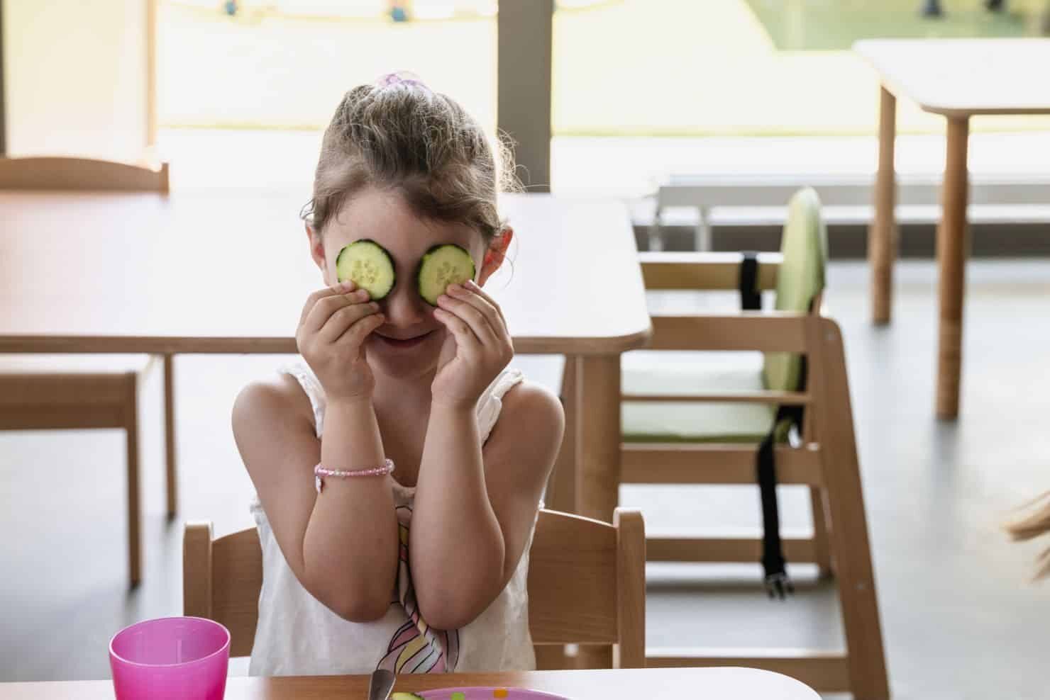 Ein kleines Mädchen sitzt an einem Tisch und hält sich Gurkenscheiben über die Augen, während im Hintergrund ein leerer Hochstuhl und andere Tische zu sehen sind. Ein kleines Mädchen sitzt an einem Tisch und hält sich Gurkenscheiben über die Augen, während im Hintergrund ein leerer Hochstuhl und andere Tische zu sehen sind.