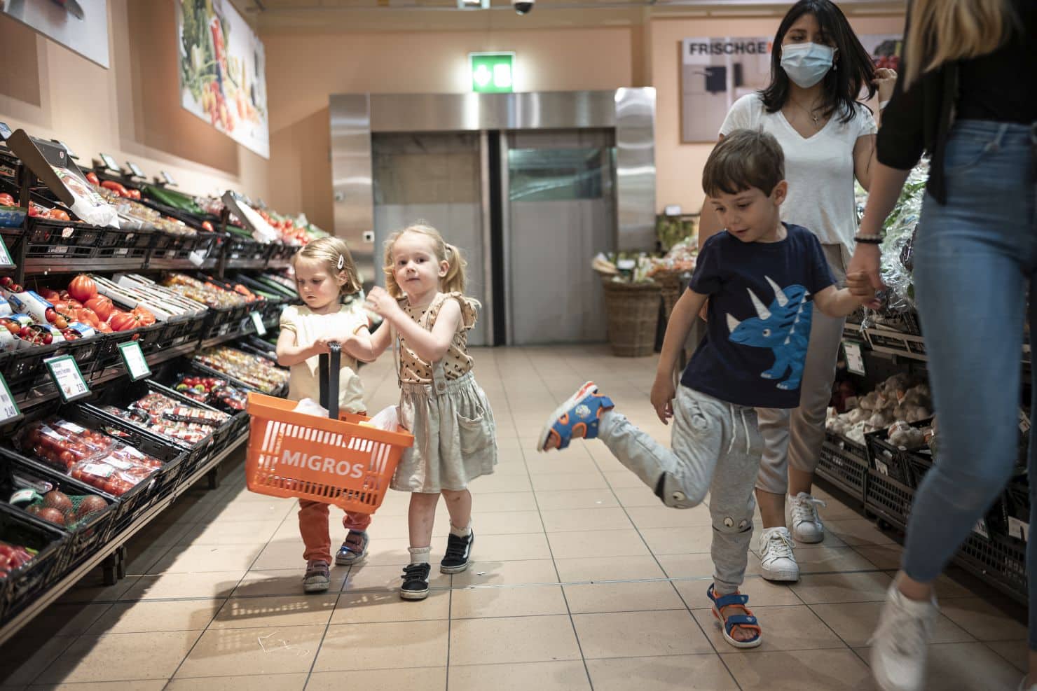Zwei kleine Mädchen tragen gemeinsam einen Migros-Einkaufskorb durch die Gemüseabteilung. Neben geht ein kleiner Junge an der Hand einer Betreuerin der Kinderkriippe miKaDo. Dahinter geht eine weitere Betreuerin.
