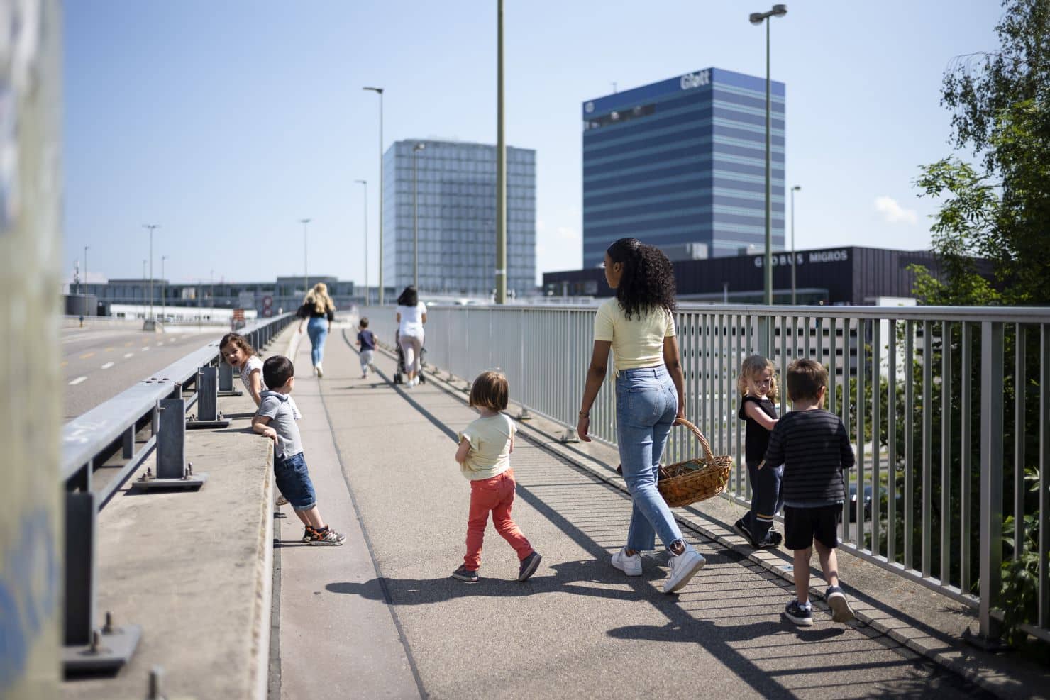 Mehrere Betreuerinnen der Kinderkrippe miKaDo gehen mit einer Kindergruppe über eine Brücke mit Geländer, hohen Gebäuden vom Glatt-Zentrum. Im Hintergrund ist klarer Himmel.