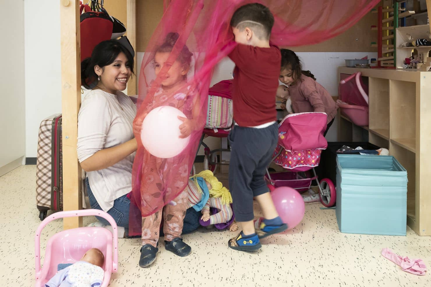 Eine Betreuerin der Kinderkrippe miKaDo und drei Kinder spielen drinnen mit rosa Luftballons und einem durchsichtigen rosa Stoff. Spielzeug, Kinderwagen und Aufbewahrungskästen sind im Raum verstreut.