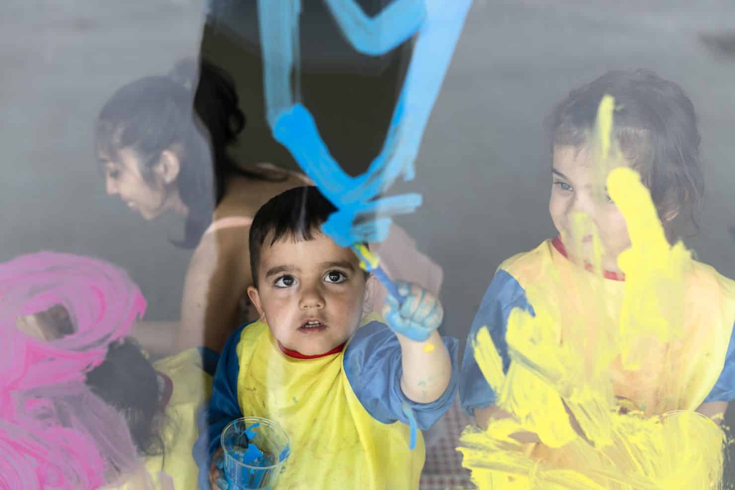 Zwei kleine Kinder in gelben Kitteln malen bunte Formen auf eine Fensterscheibe, wobei blaue, gelbe und rosa Farbe zu sehen ist. Im Hintergrund sieht man eine Betreuerin der Kinderkrippe miKaDo, die sich lächelnd anderen Kindern widmet.