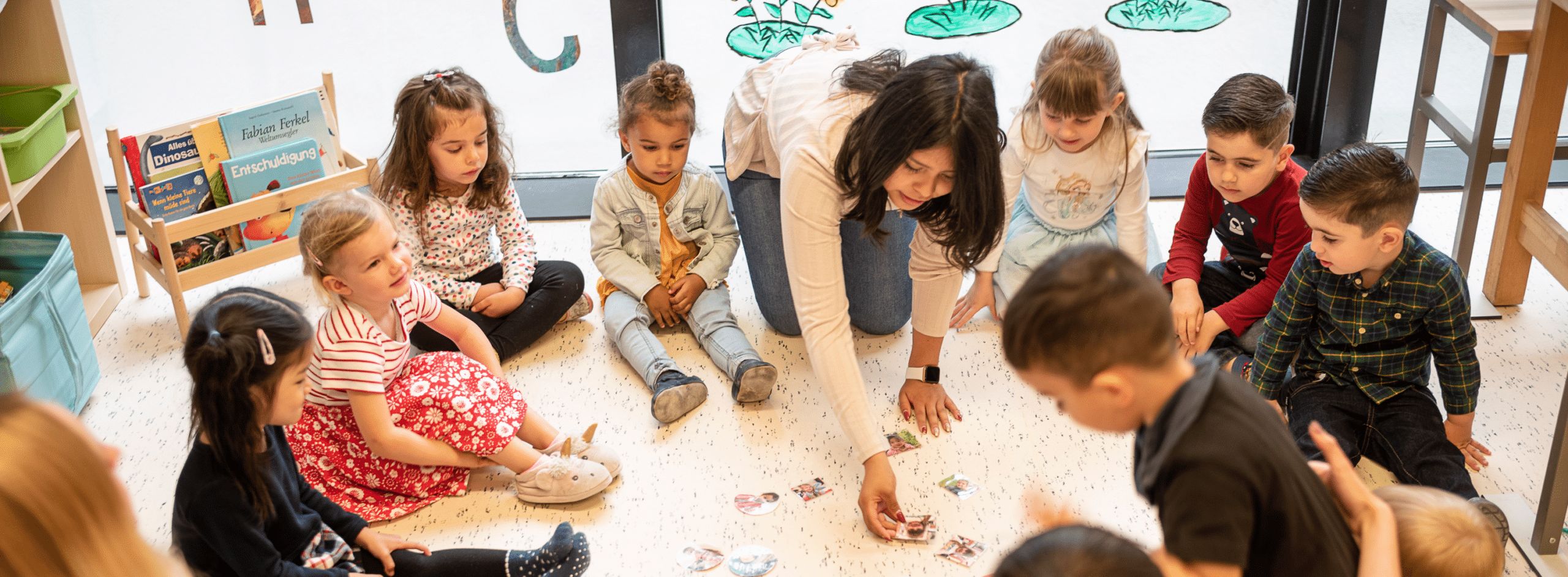 Eine Lehrerin und eine Gruppe kleiner Kinder sitzen im Kreis auf dem Boden und beschäftigen sich während einer Unterrichtsaktivität mit Bildkarten.